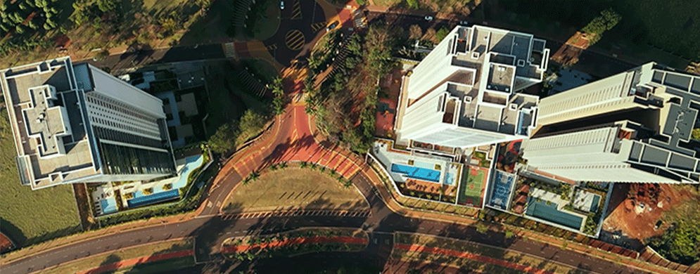 Vista aérea mostra dois prédios modernos com piscinas e ruas arborizadas ligadas por rotatória central
