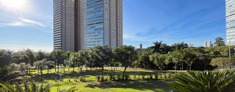 Jardim arborizado com gramado e palmeiras em primeiro plano, tendo ao fundo um alto edifício residencial moderno