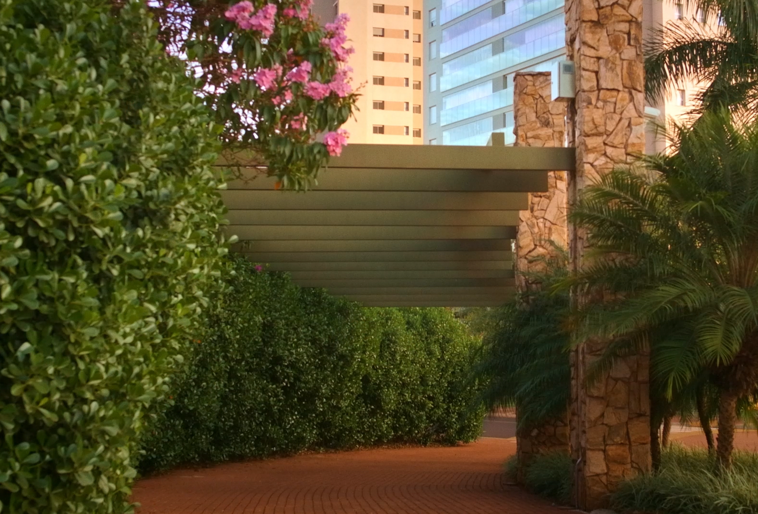 Caminho arborizado com pergolado de metal verde, colunas de pedra e prédios modernos ao fundo.