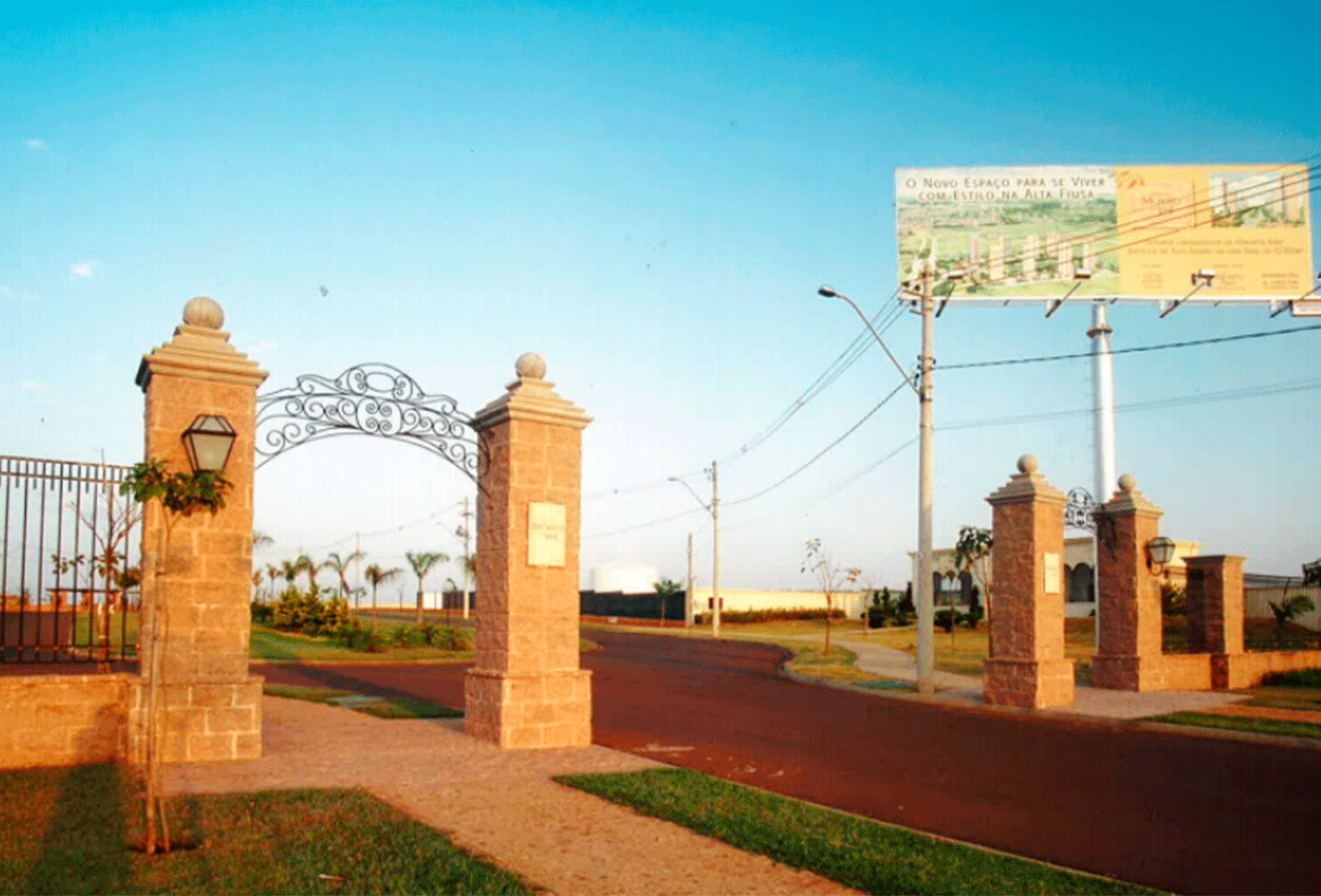 Entrada de condomínio com portais de pedra, portão de ferro e arco decorativo, ao lado de avenida asfaltada com outdoor