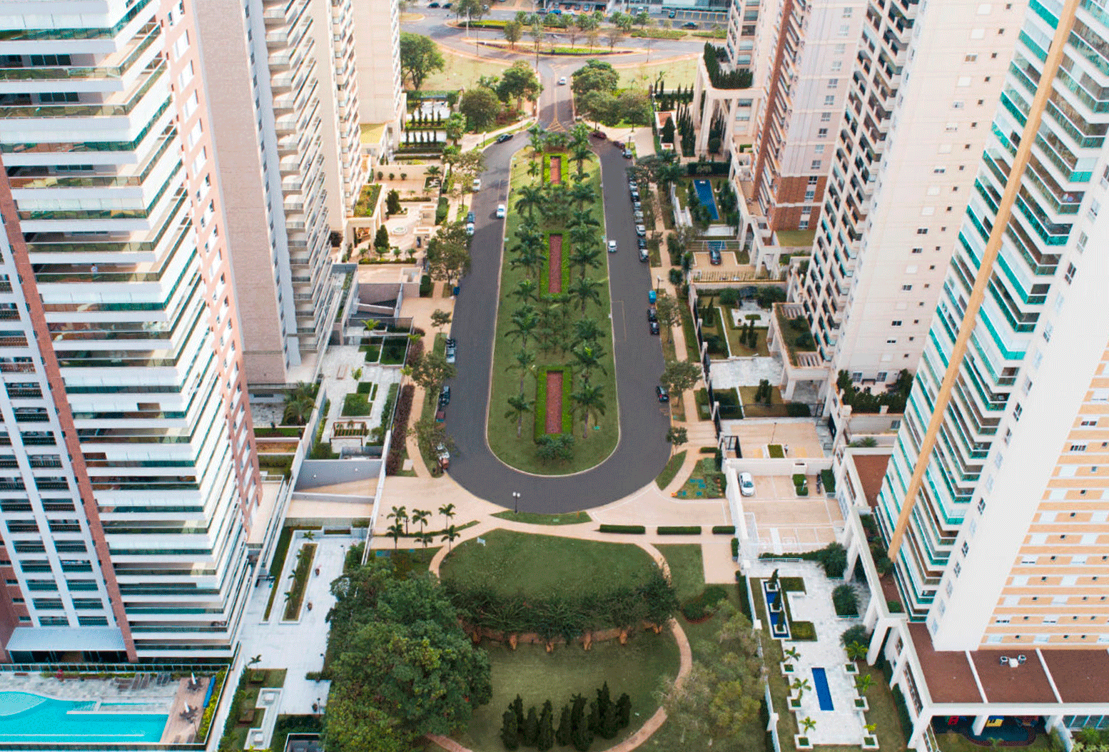 Vista aérea de avenida central arborizada cercada por altos prédios residenciais, com piscinas, jardins e áreas de lazer integradas.
