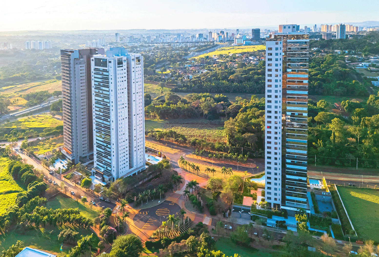 Vista aérea de três torres residenciais de alto padrão, cercadas por áreas verdes, vias arborizadas e a cidade ao fundo.