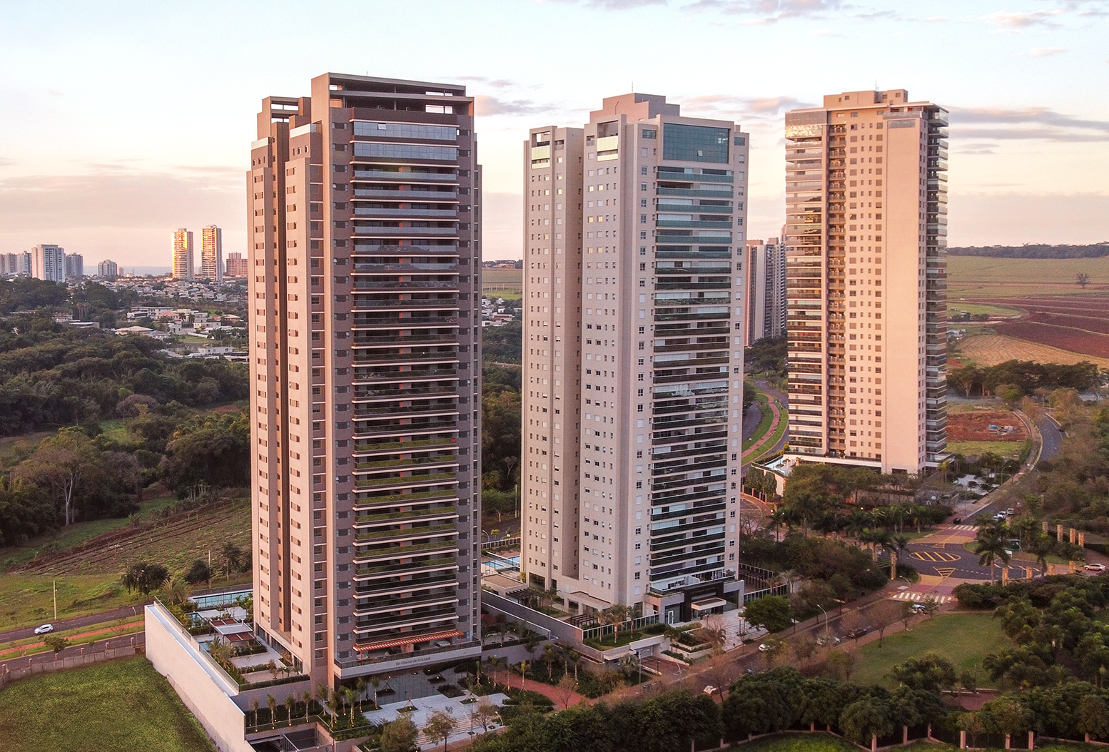 Vista aérea de três prédios altos e modernos, cercados por ruas, áreas verdes e a cidade ao fundo.