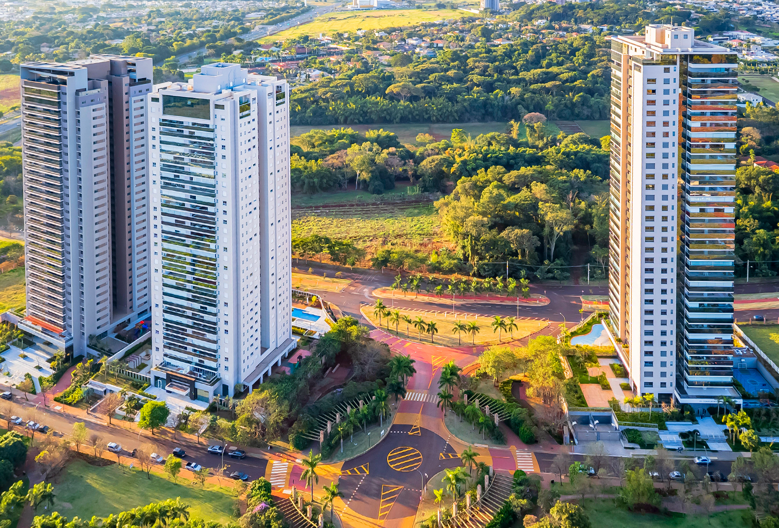 Vista aérea de torres residenciais altas, ruas arborizadas, áreas verdes e cidade ao fundo.