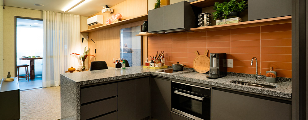 Cozinha compacta com armários cinza, bancada de granito, revestimento terracota e sala ao fundo.