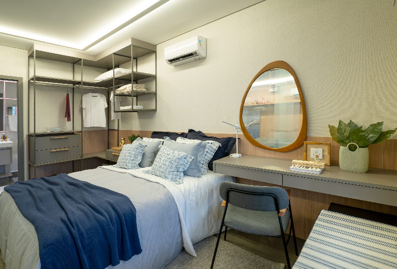 Quarto com cama de casal, closet aberto, escrivaninha com espelho redondo e banheiro ao fundo.