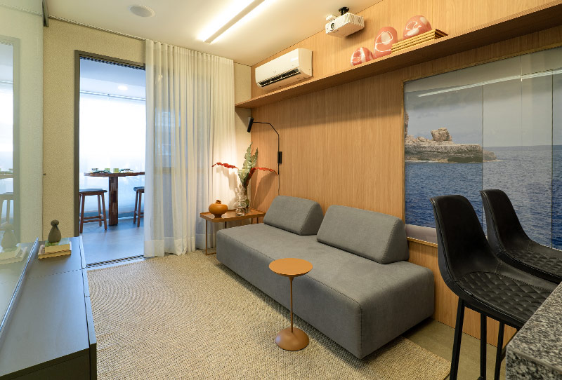 Sala compacta com sofá cinza, painel de madeira, banquetas e varanda iluminada ao fundo.