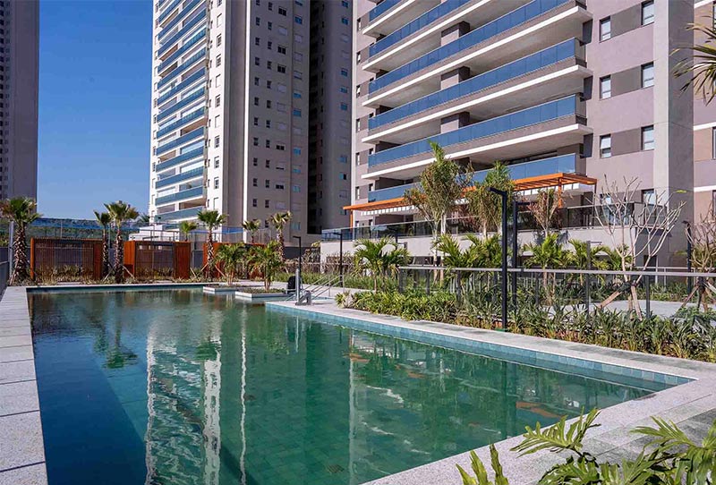 Imagem com grande piscina em primeiro plano, vegetação vasta ao redor e torres imponentes ao fundo com varandas grandes.