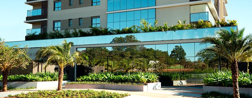 Fachada de edifício moderno com vidros espelhados, jardins, palmeiras e entrada ampla junto à calçada.