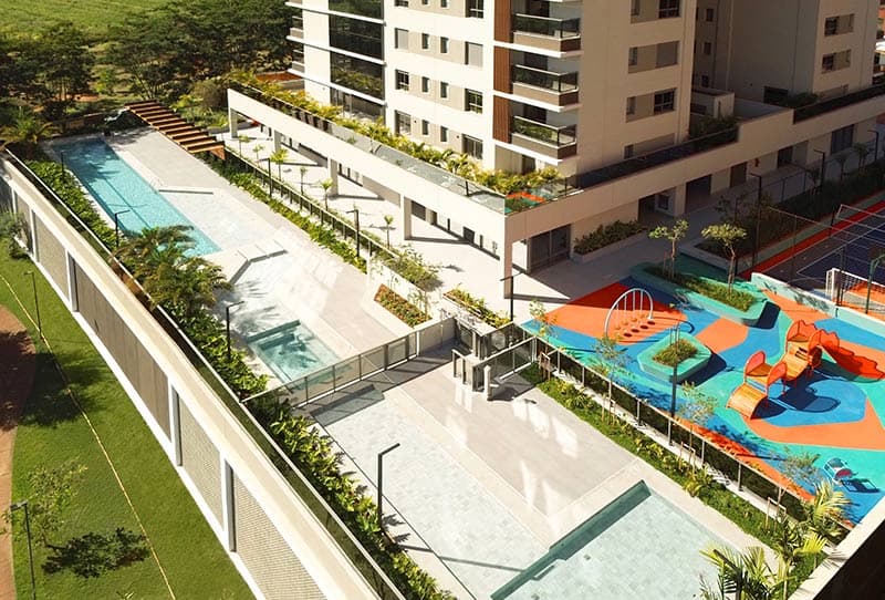 Área de lazer vista do alto, com piscinas, playground colorido, quadra esportiva e jardins ao redor.