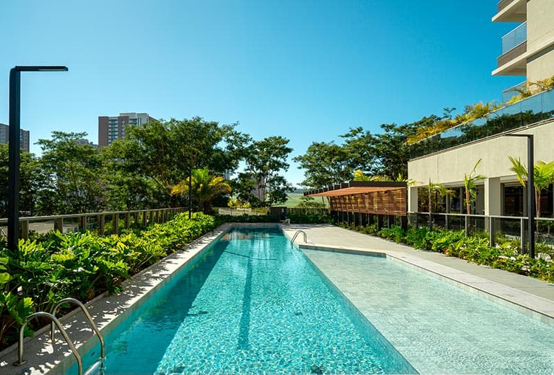 Piscina adulto ao ar livre junto a edifício alto, cercada por jardins e áreas de convivência.