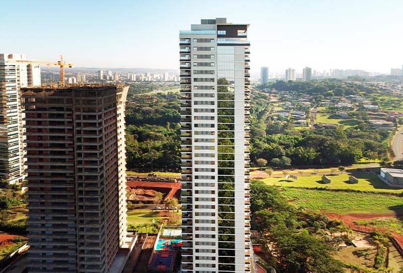 Vista aérea de edifício residencial alto, com obra ao lado, áreas verdes e horizonte urbano ao fundo.